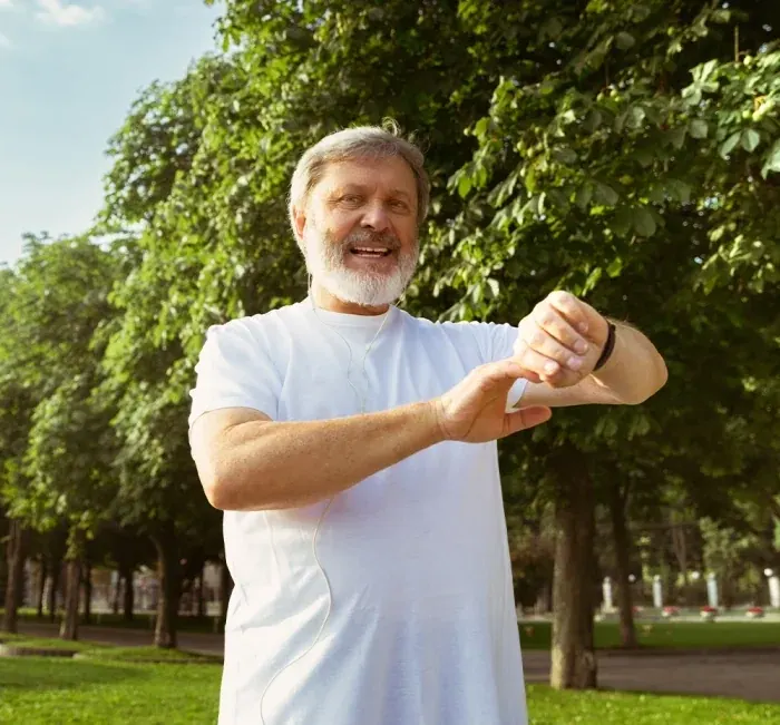 A smiling older man in a white T-shirt checking his watch in a park